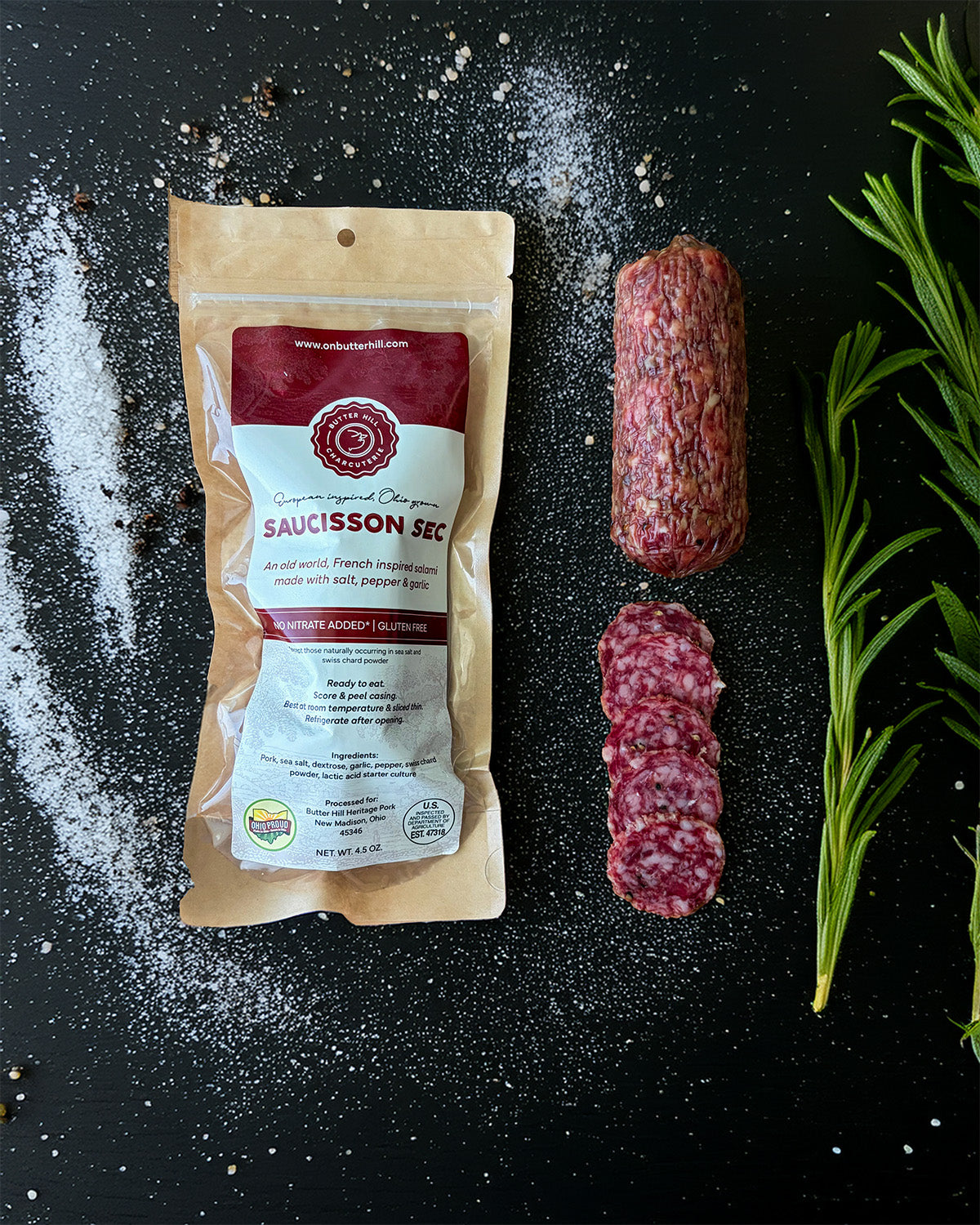 Sauccisson sec packaging with sliced salami and rosemary on a black surface
