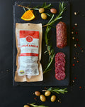 Loukanika package with sliced salami on a dark slate board with herbs and olives.