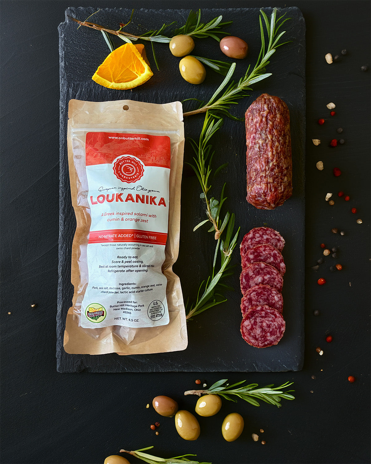 Loukanika package with sliced salami on a dark slate board with herbs and olives.